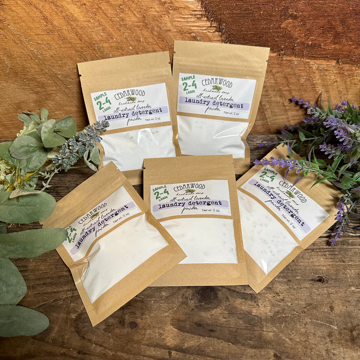 Five packages of laundry detergent with labels on a wooden surface, surrounded by greenery and flowers.